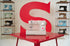 A beige Singer sewing machine sits on a glossy red table, with a large red letter S on the wall behind it and shelves of vintage sewing machines on both sides.