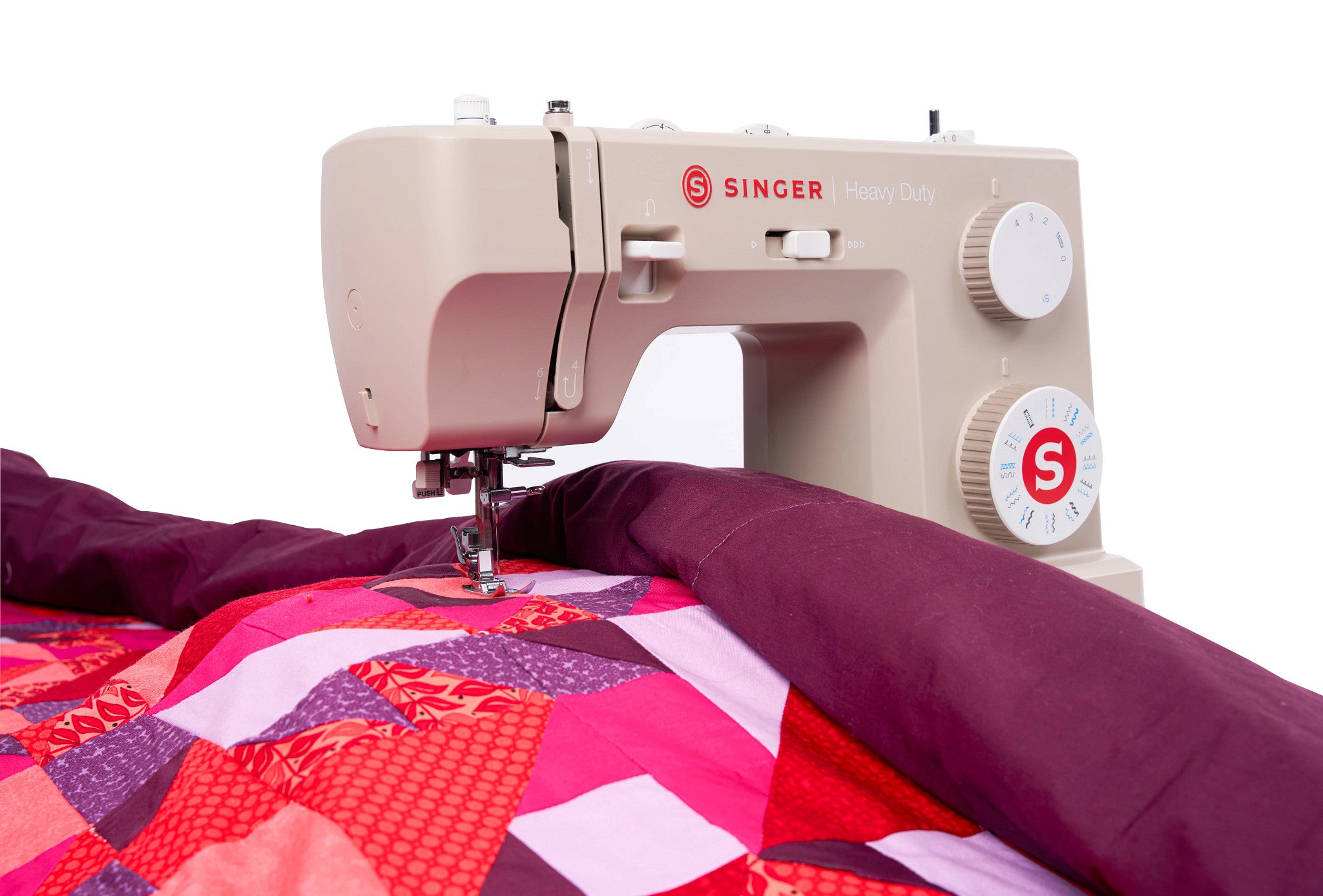 A beige Singer Hilary Daily sewing machine actively sewing a colorful patchwork quilt with a deep purple fabric layer, shown against a solid white background.
