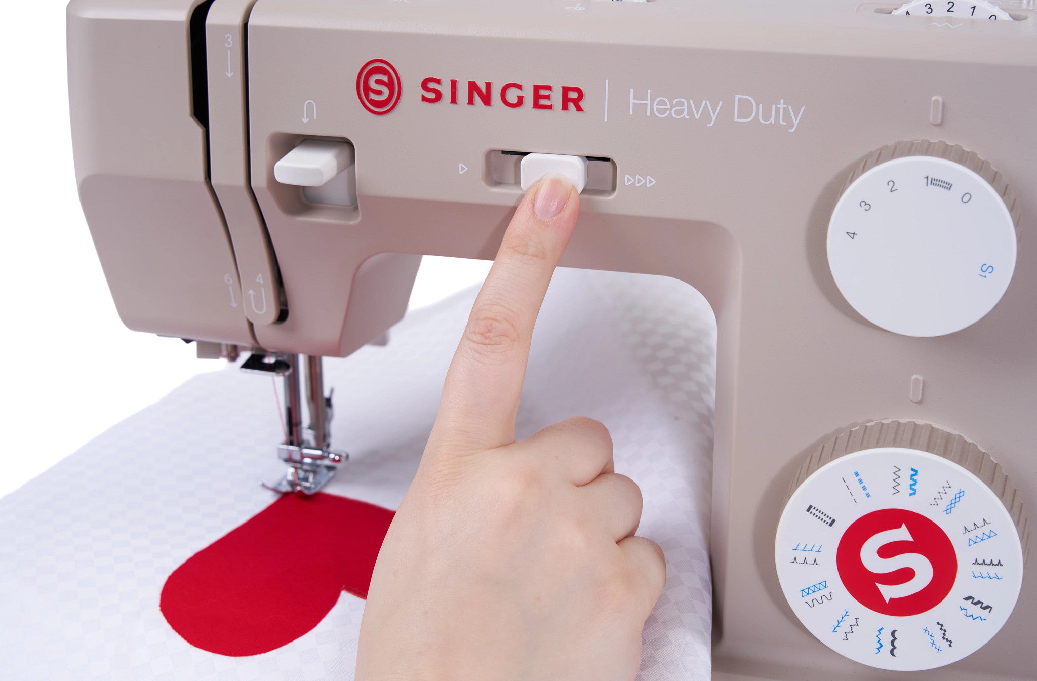 A close-up of a beige Singer Heavy Duty sewing machine with a person's finger pressing a button near the needle area, with white fabric and a red heart-shaped design under the press.