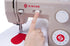 A close-up of a beige Singer Heavy Duty sewing machine with a person's finger pressing a button near the needle area, with white fabric and a red heart-shaped design under the press.