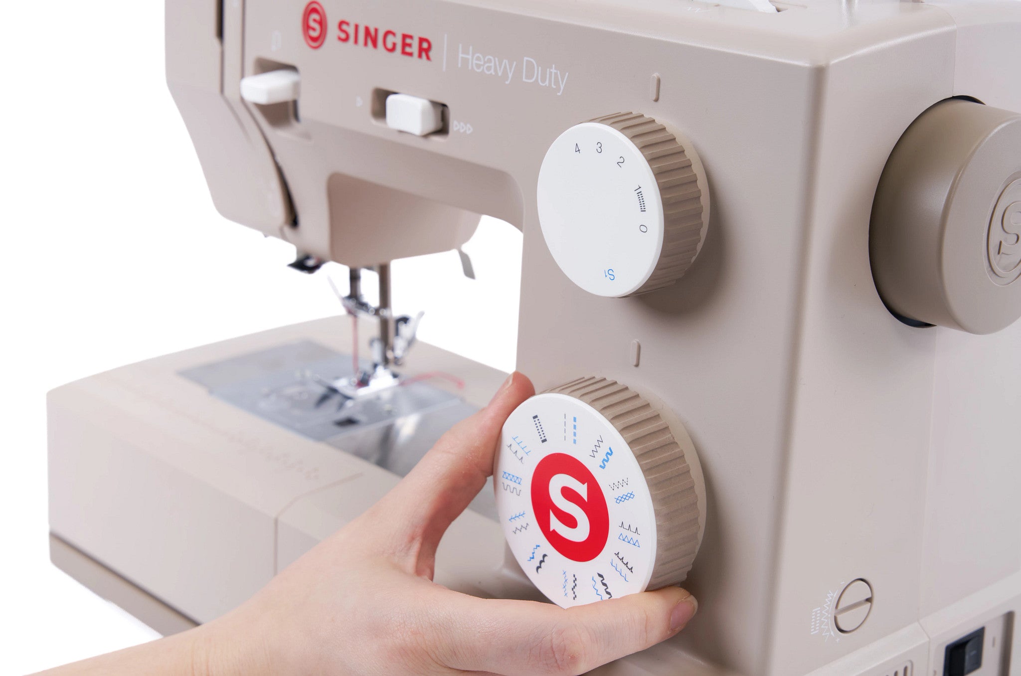 A close-up of a beige Singer Heavy Duty sewing machine with a hand turning one of the large control dials. The machine features a white dial with a red 'S' logo and blue stitch settings, and another white dial with red 'S' branding.