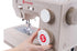 A close-up of a beige Singer Heavy Duty sewing machine with a hand turning one of the large control dials. The machine features a white dial with a red 'S' logo and blue stitch settings, and another white dial with red 'S' branding.