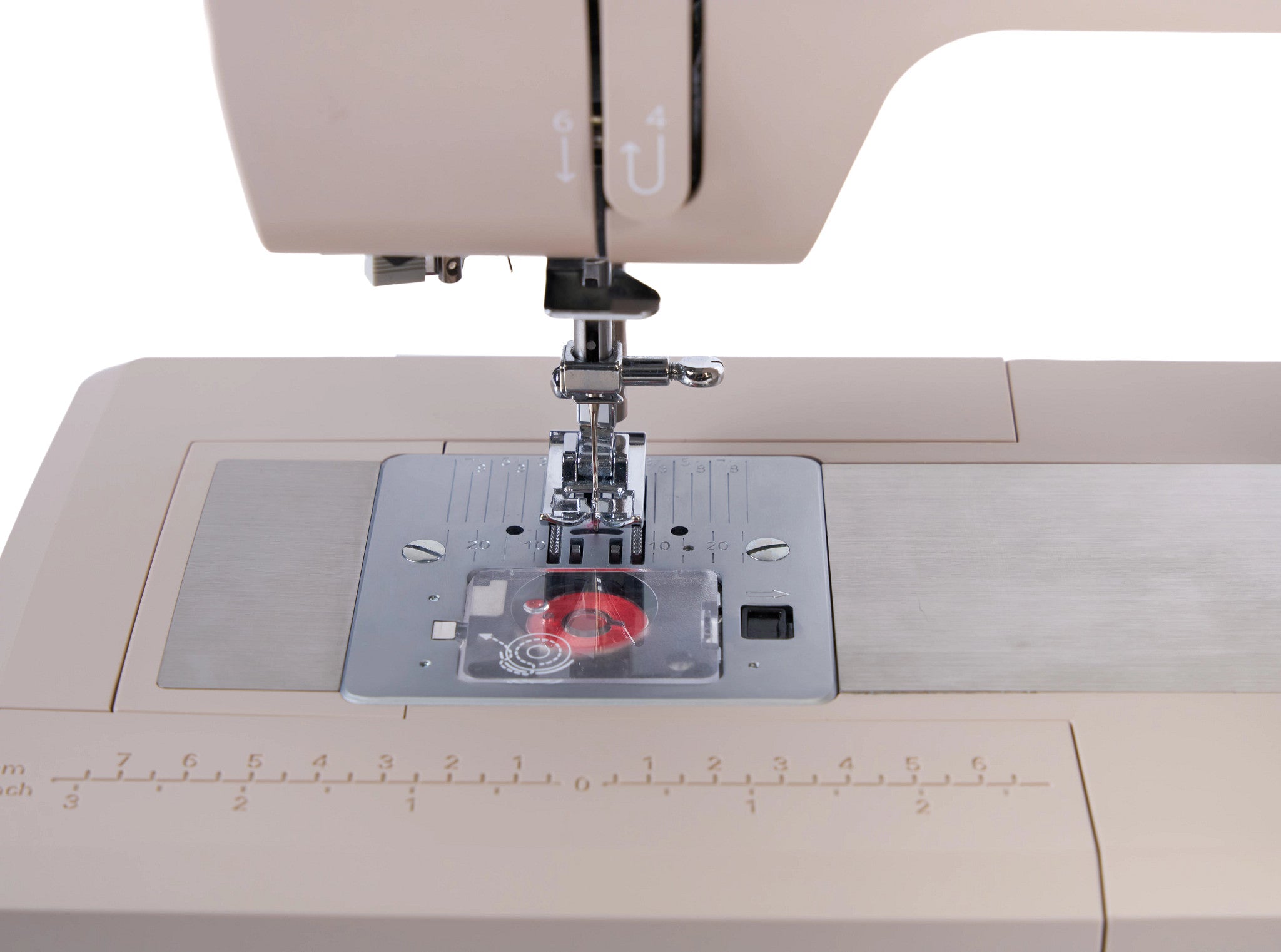 Close-up of a sewing machine needle area with a beige machine body, silver metal plate, clear cover, and a red spool of thread on a white background.