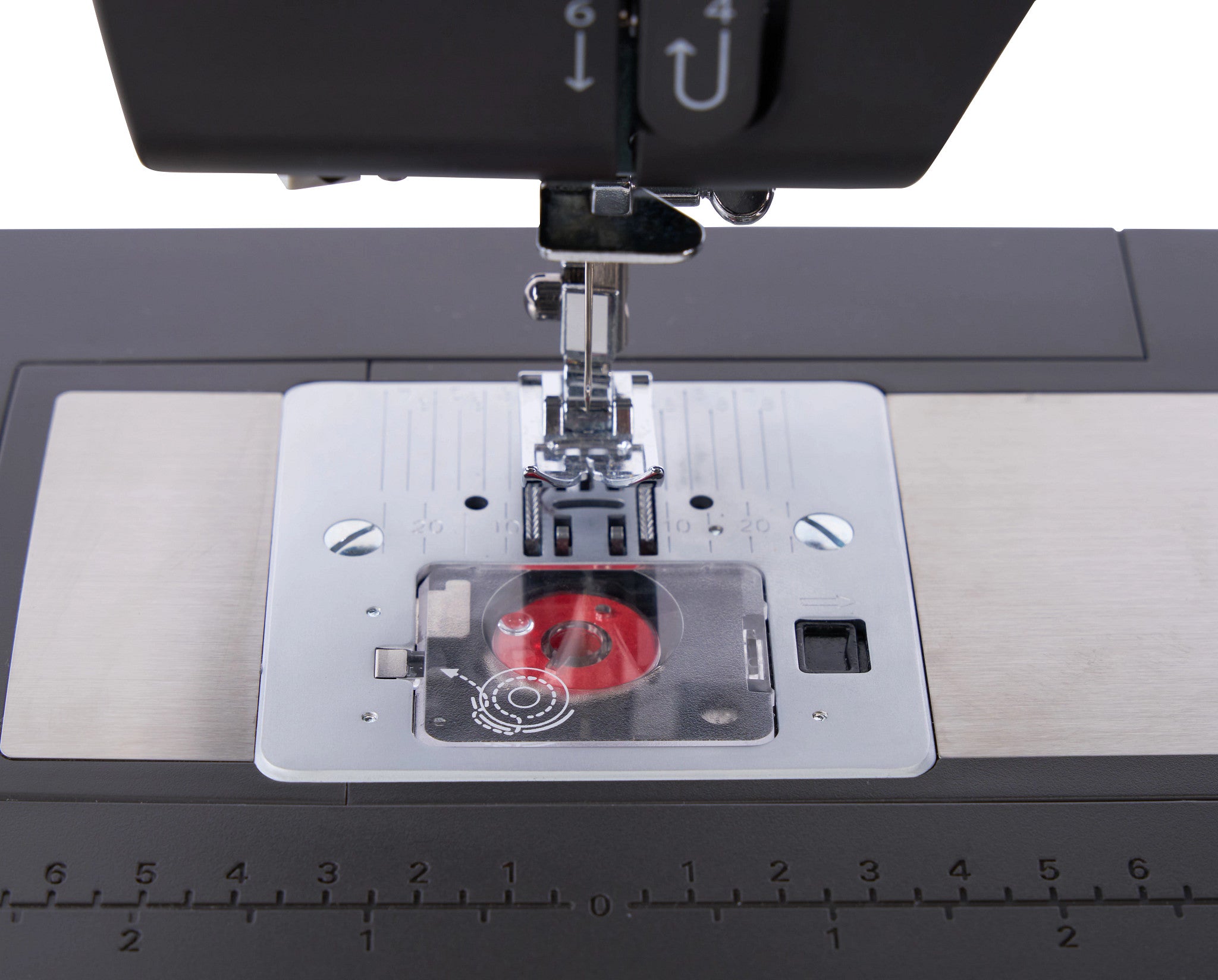Close-up of a sewing machine needle plate area with a clear cover over a red spool of thread, mounted on a dark gray machine bed with a silver metal plate and ruler markings.