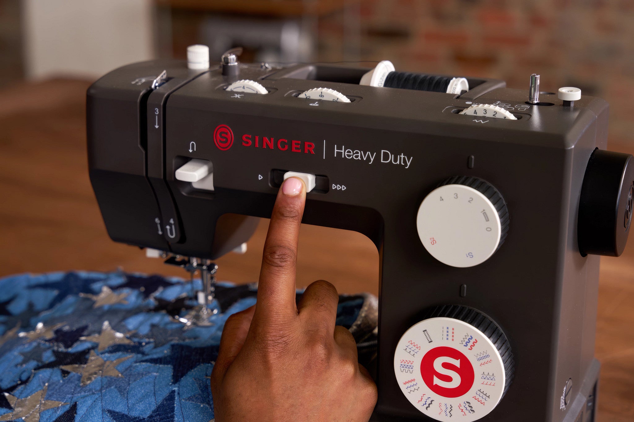 A close-up of a black Singer Heavy Duty sewing machine with a person's finger pressing a button, set against a warm indoor background with a blue patterned fabric underneath.
