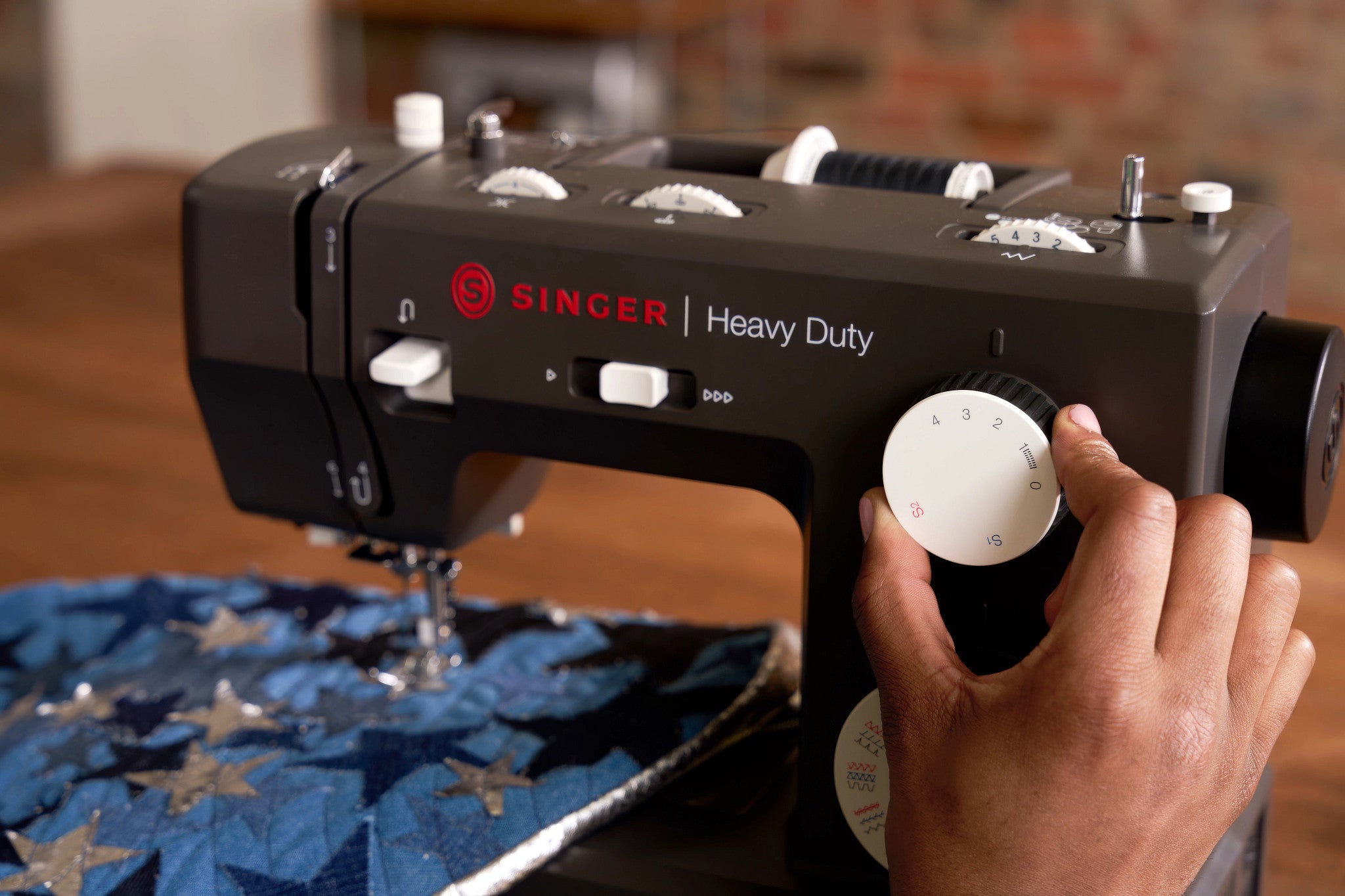 A close-up of a black Singer Heavy Duty sewing machine being operated by a person's hand, with blue fabric featuring silver star patterns under the needle.