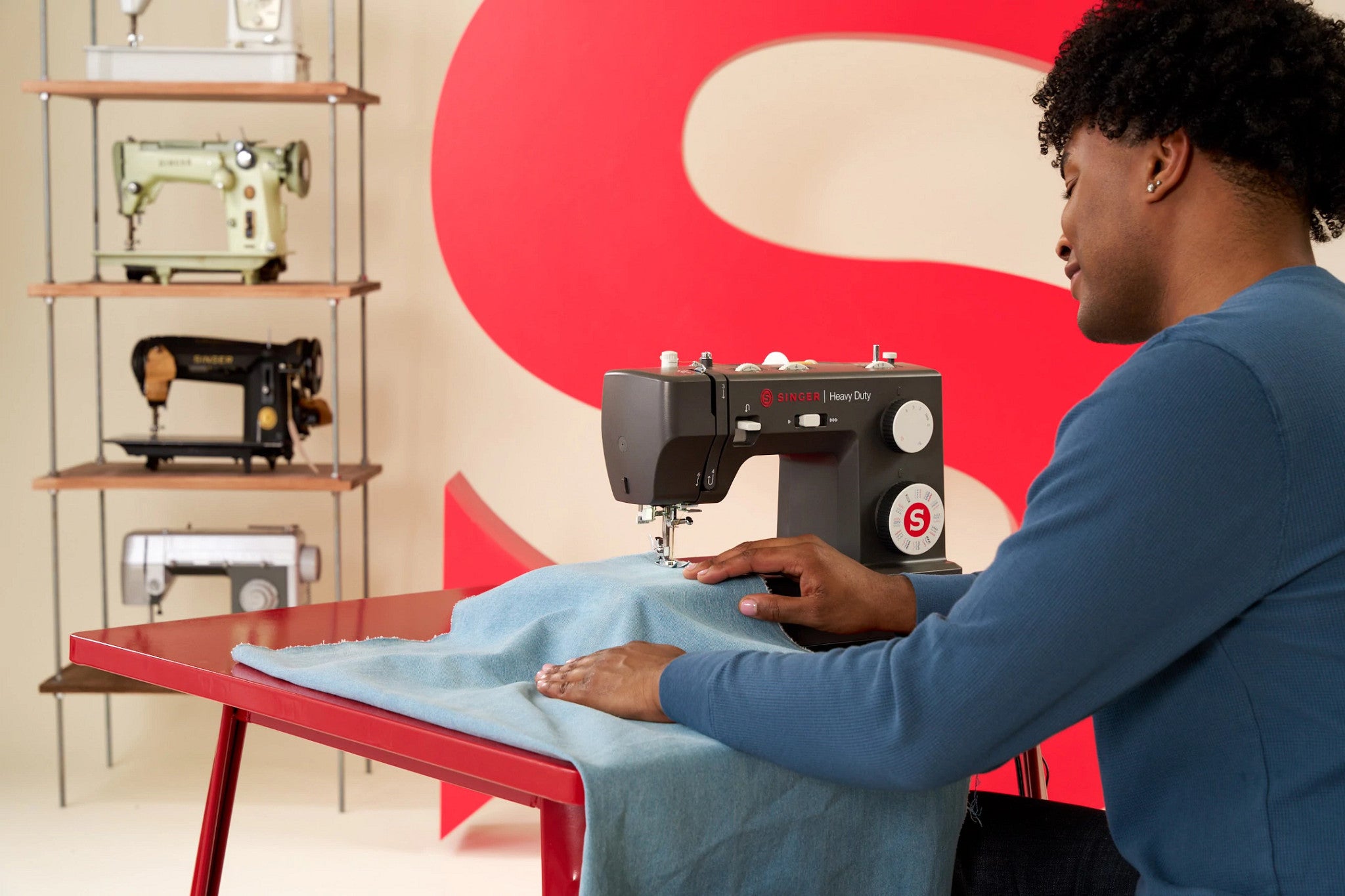 A person in a blue long-sleeve shirt operates a black and white Brother sewing machine, guiding light blue fabric under the needle. The scene is set in a studio with a red table, a large red background graphic, and shelves displaying other vintage sewing machines.
