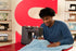 A man in a blue henley shirt operates a dark gray Singer sewing machine, guiding a light blue fabric under the needle. The background features a large red 'S' logo on a beige wall with shelves displaying other vintage sewing machines.