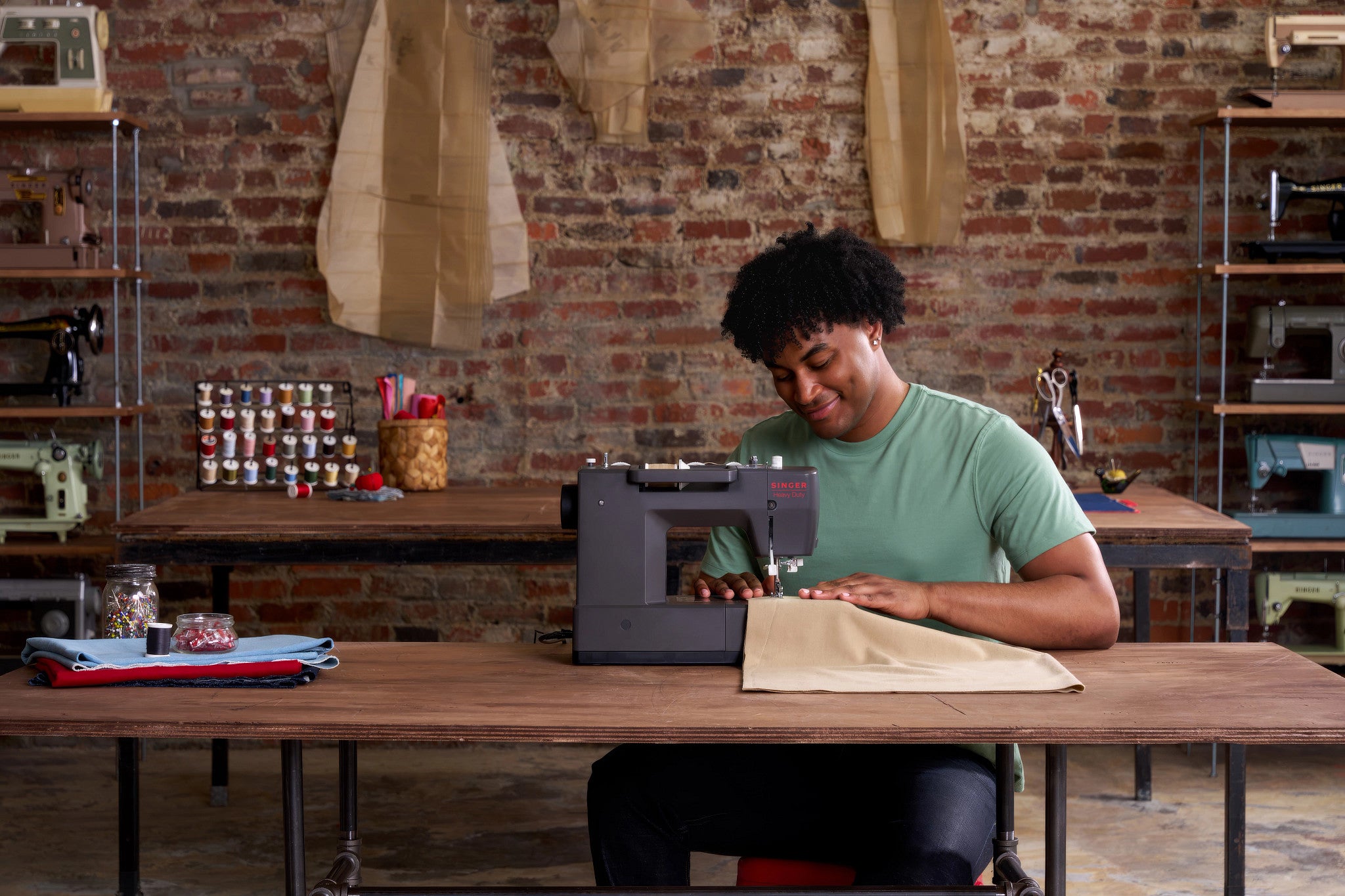 A person sitting at a wooden table in a workshop, using a gray sewing machine with beige fabric. The background features a rustic brick wall, shelves with sewing equipment, and hanging fabric pieces.