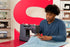 A man in a blue henley shirt operates a Singer sewing machine, guiding light blue fabric under the needle. The scene is set in a workshop with shelves of vintage sewing machines and a large red circular backdrop.