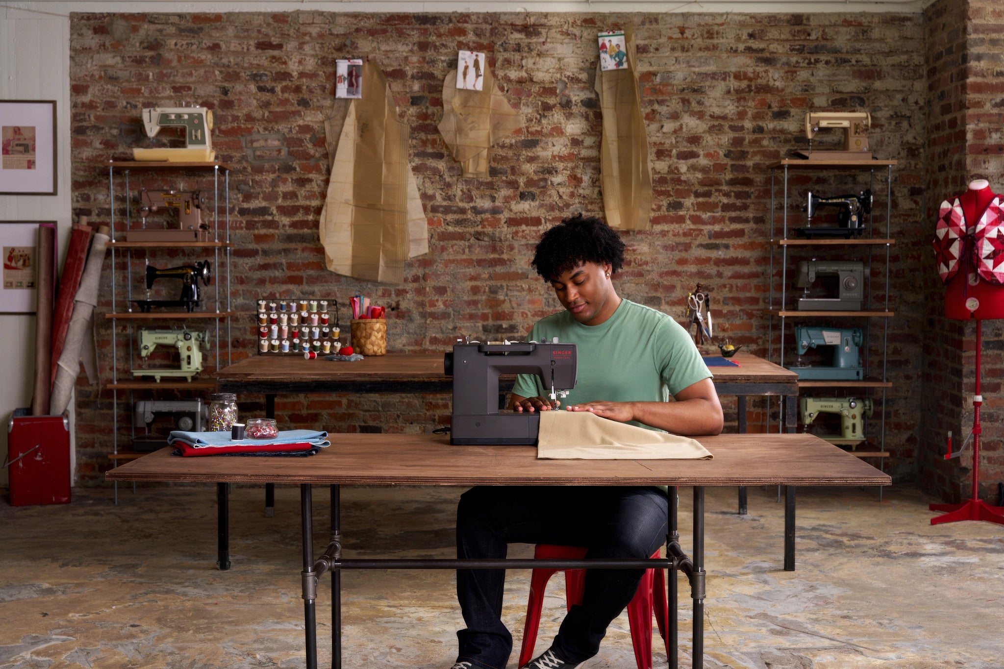 A person sits at a wooden table in a rustic workshop with exposed brick walls, sewing machines, and hanging fabric samples, using a sewing machine with a piece of beige fabric.