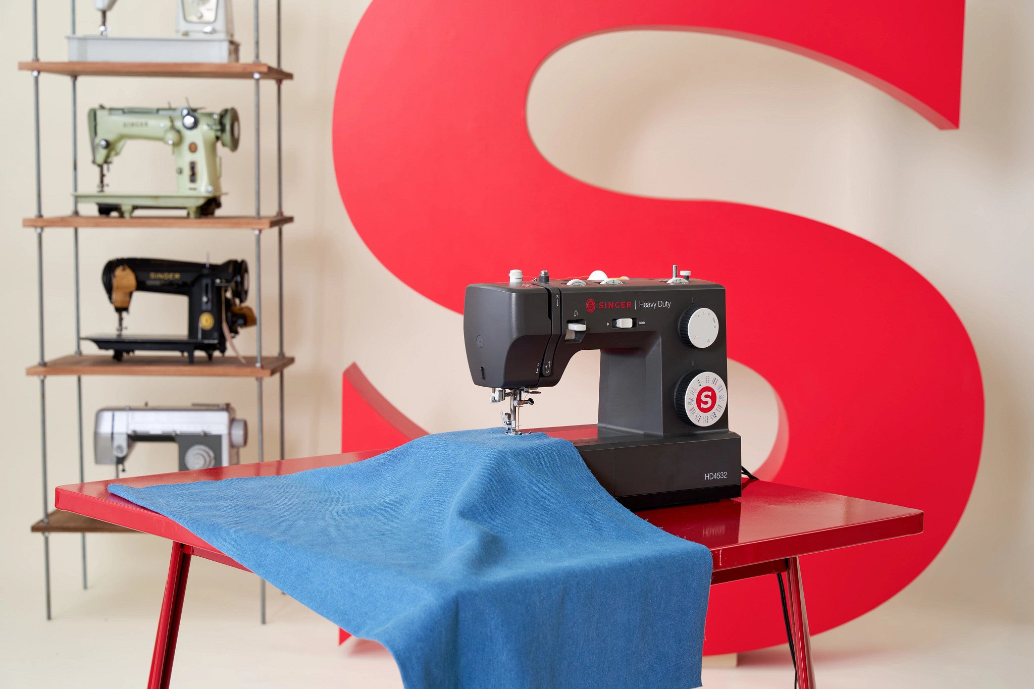 A black Brother sewing machine sits on a red table draped with blue fabric, with a large red letter S on the wall behind it. Shelves with other sewing machines are visible on the left.