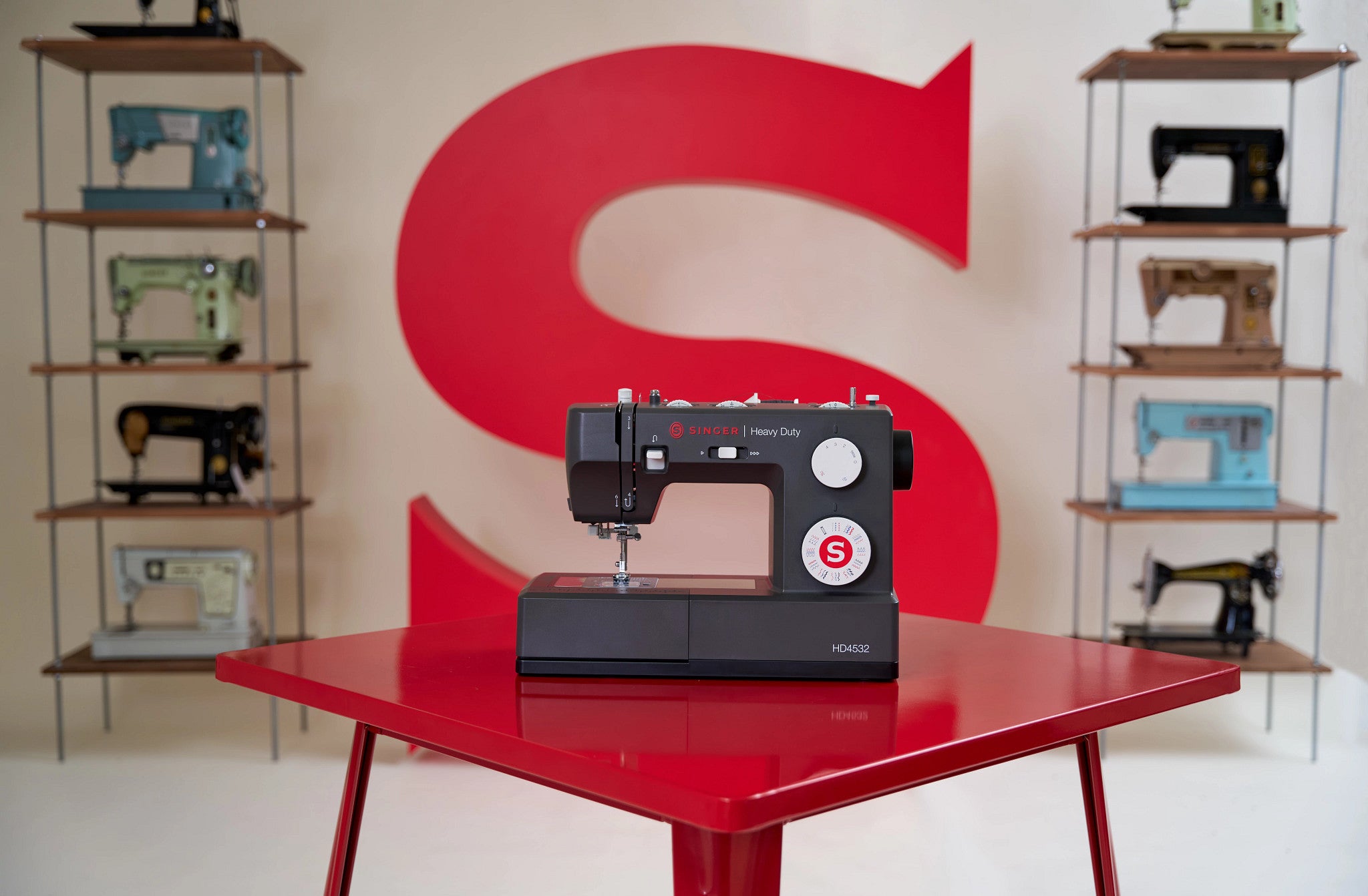 A black Singer sewing machine sits on a glossy red table, with a large red letter S on the wall behind it and shelves of other vintage sewing machines on both sides.