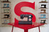 A black Singer sewing machine sits on a glossy red table, with a large red letter S on the wall behind it and shelves of other vintage sewing machines on both sides.