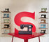 A black and red Singer sewing machine sits on a red table in the center, with a large red 'S' logo on the wall behind it. Shelves on both sides display other vintage sewing machines.