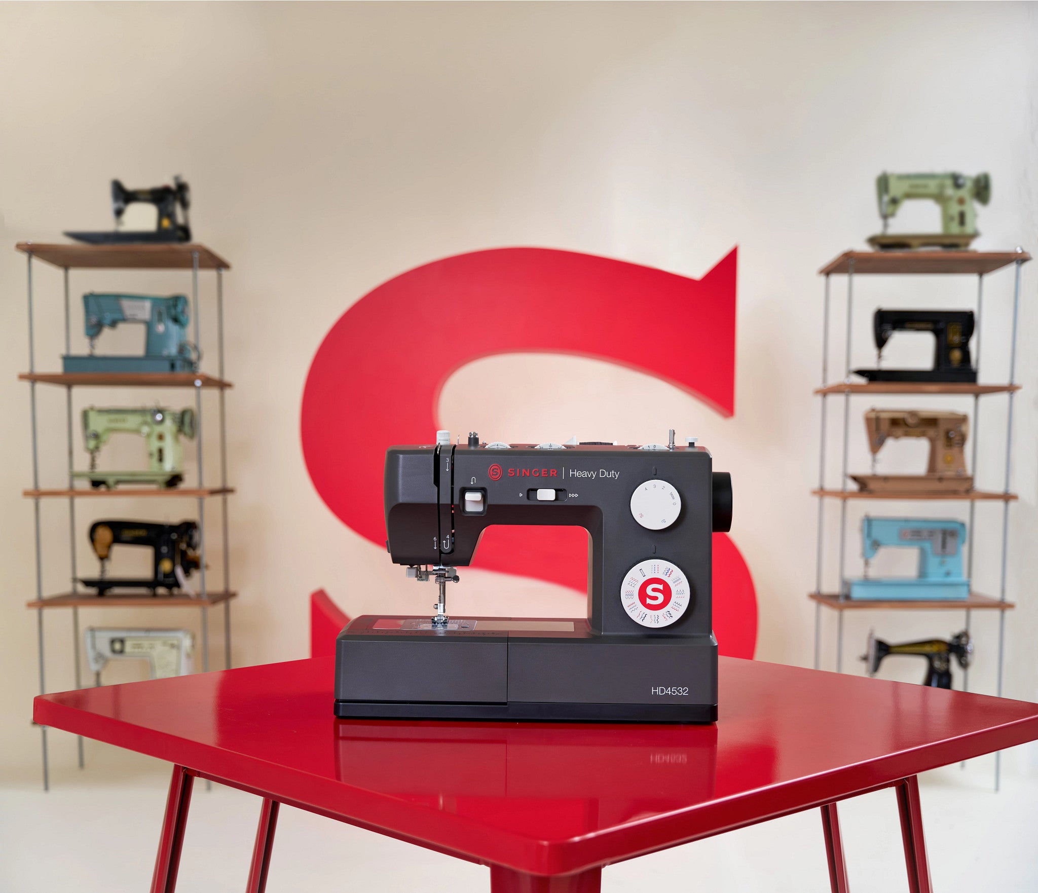 A black Singer sewing machine sits on a glossy red table, with a large red 'S' logo on the wall behind it and vintage sewing machines displayed on shelving units on both sides.