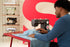 A person in a blue long-sleeve shirt operates a black and white Brother sewing machine, guiding light blue fabric under the needle. The scene is set in a studio with a red table, a large red background graphic, and shelves displaying other vintage sewing machines.