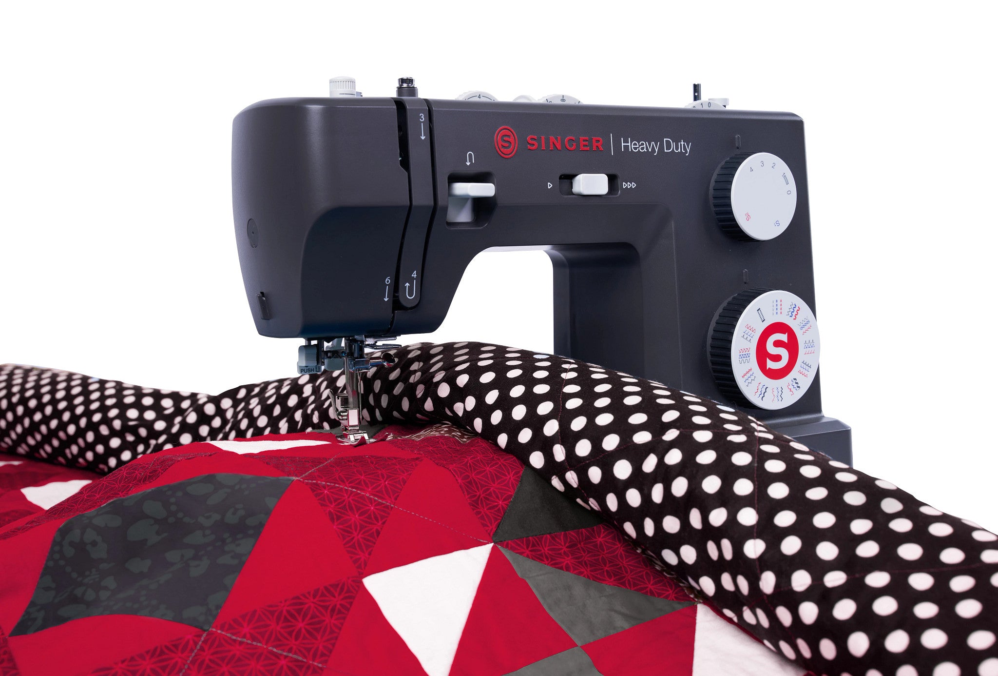 A black Singer Heavy Duty sewing machine actively stitching a red, black, and white geometric quilt, shown against a clean white background.