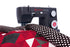 A black Singer Heavy Duty sewing machine actively stitching a red, black, and white geometric quilt, shown against a clean white background.