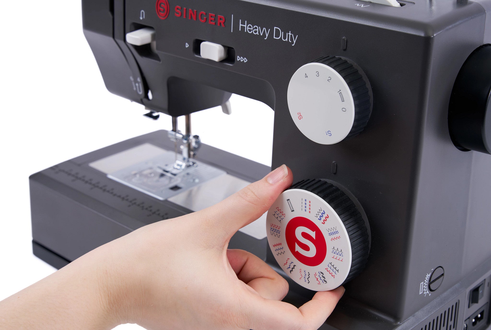 A close-up of a black Singer Heavy Duty sewing machine with a person's hand adjusting a white and black speed control dial. The brand name 'SINGER' and 'Heavy Duty' are printed on the machine in red and white.