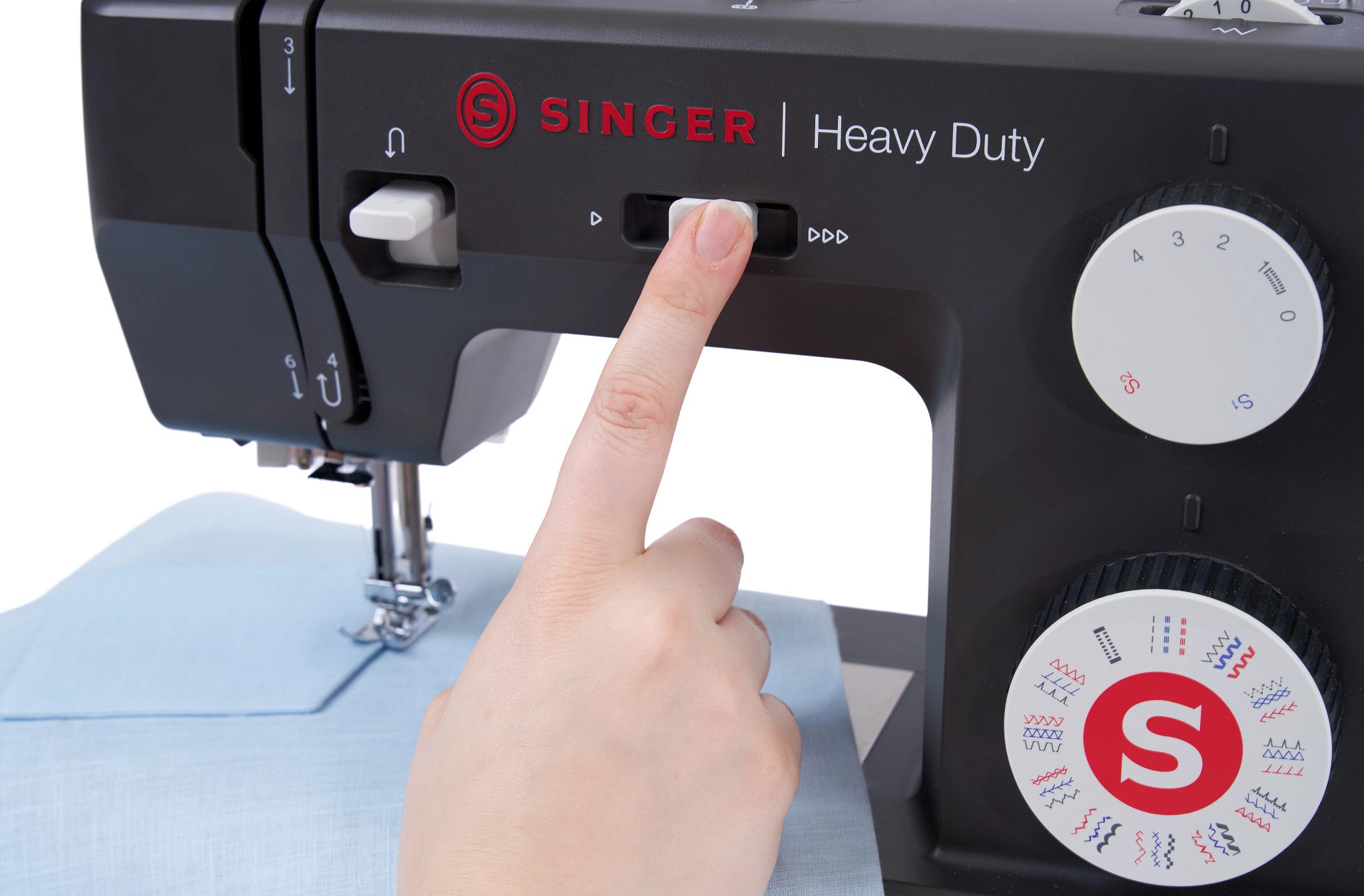 A close-up of a black Singer Heavy Duty sewing machine with a person's finger pressing a button, with a light blue fabric under the needle.
