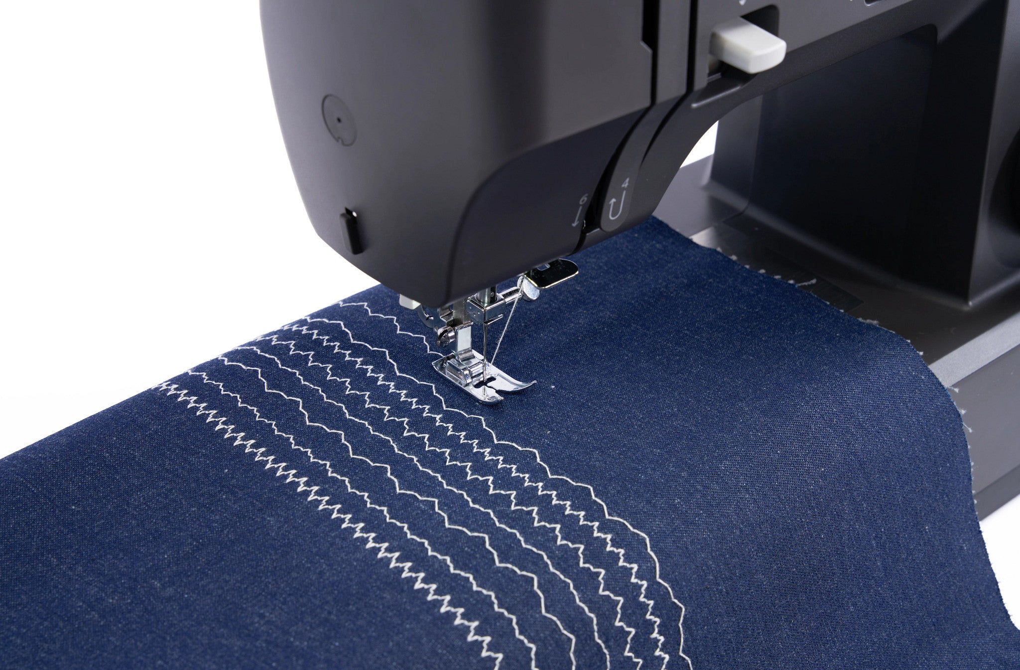 Close-up of a black sewing machine stitching dark blue denim fabric with white zigzag and straight stitch details on a white background.
