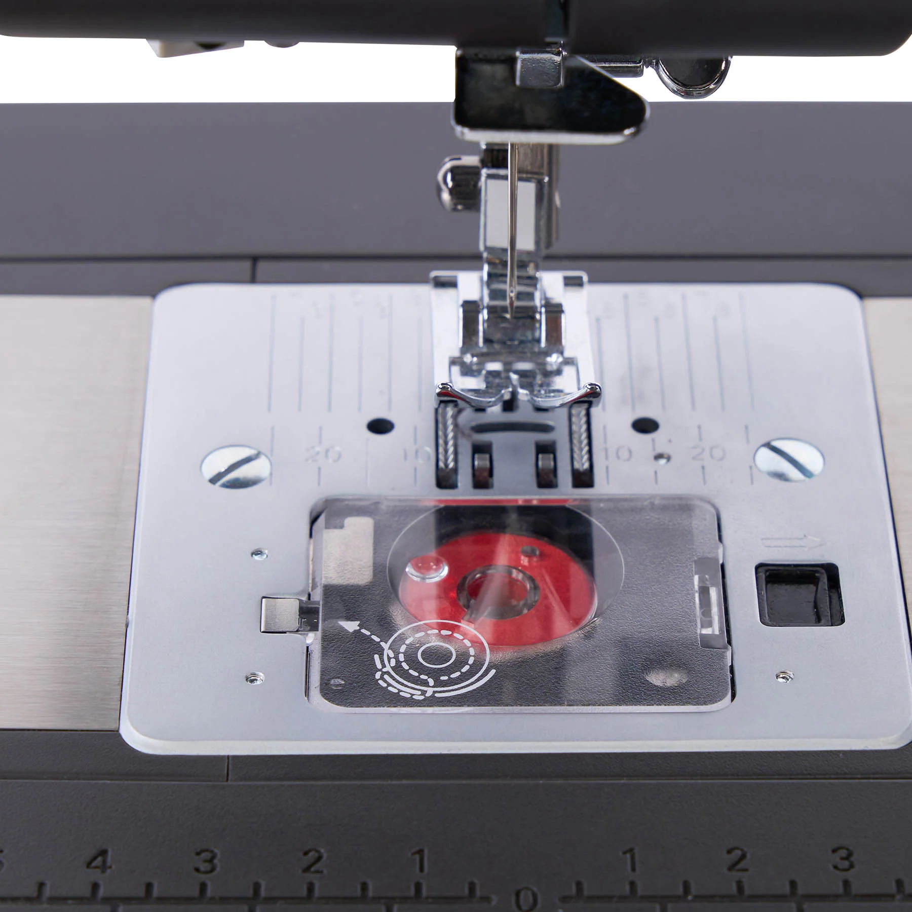 Close-up of a sewing machine needle plate area with a silver metal plate, black presser foot, and a red spool holder cover inside a clear plastic guard.