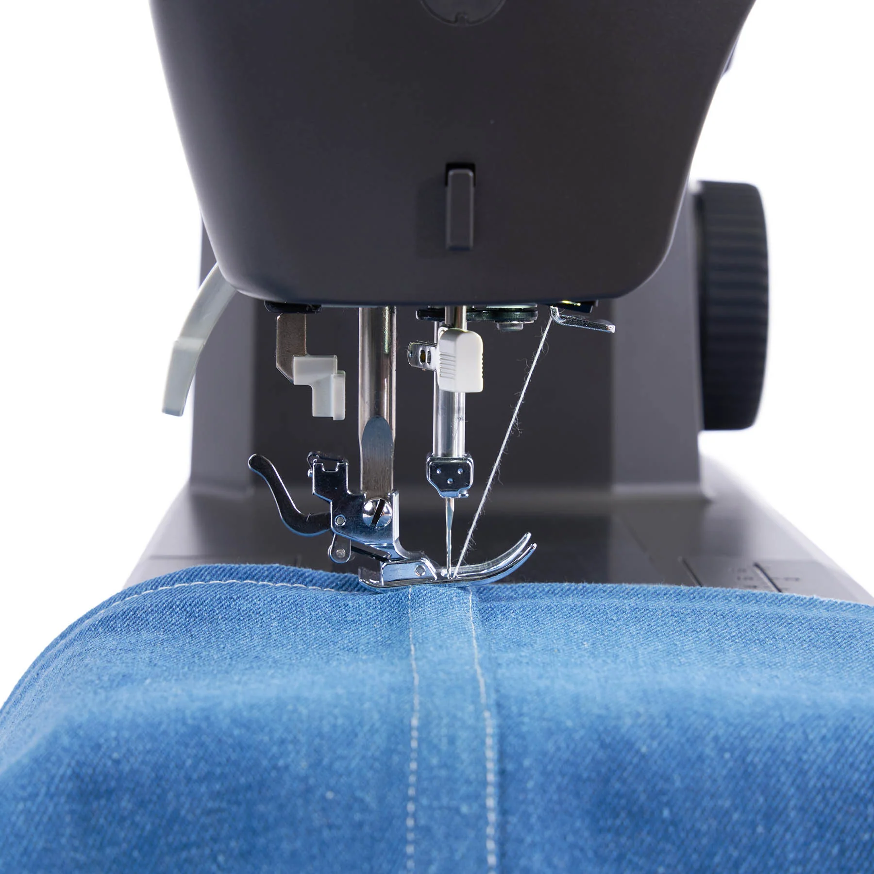 Close-up of a sewing machine needle and presser foot stitching blue denim fabric, with the black sewing machine body visible above and a white background around the edges.