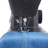 Close-up of a sewing machine needle and presser foot stitching blue denim fabric, with the black sewing machine body visible above and a white background around the edges.