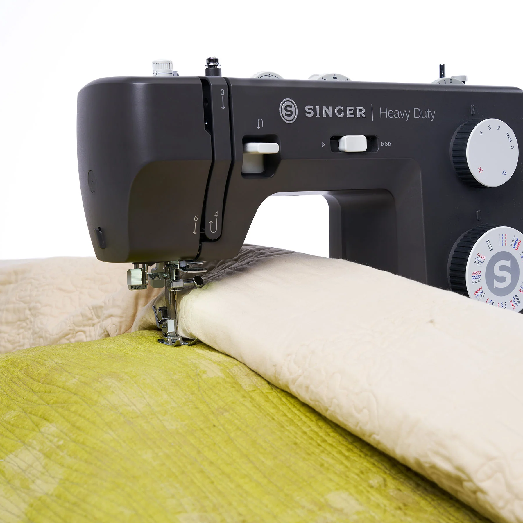 A close-up of a black Singer Heavy Duty sewing machine stitching a light beige quilt over a lime green fabric, set against a clean white background.