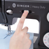 A close-up of a hand pressing a button on a black Singer Heavy Duty sewing machine, with a light blue fabric visible underneath the needle area.