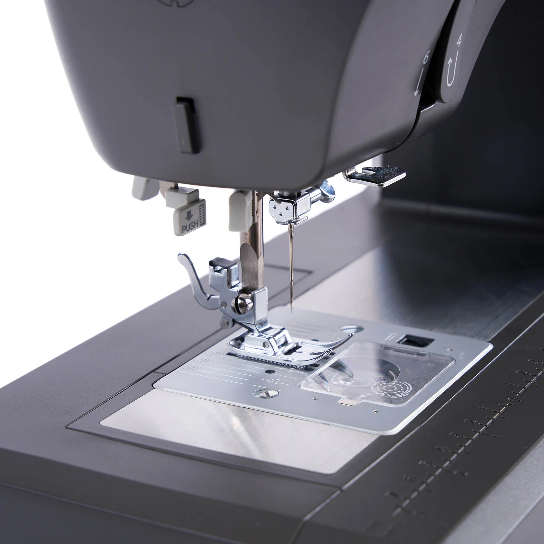 Close-up of a dark gray sewing machine showing the presser foot, needle, feed dog, and metallic needle plate against a white background.