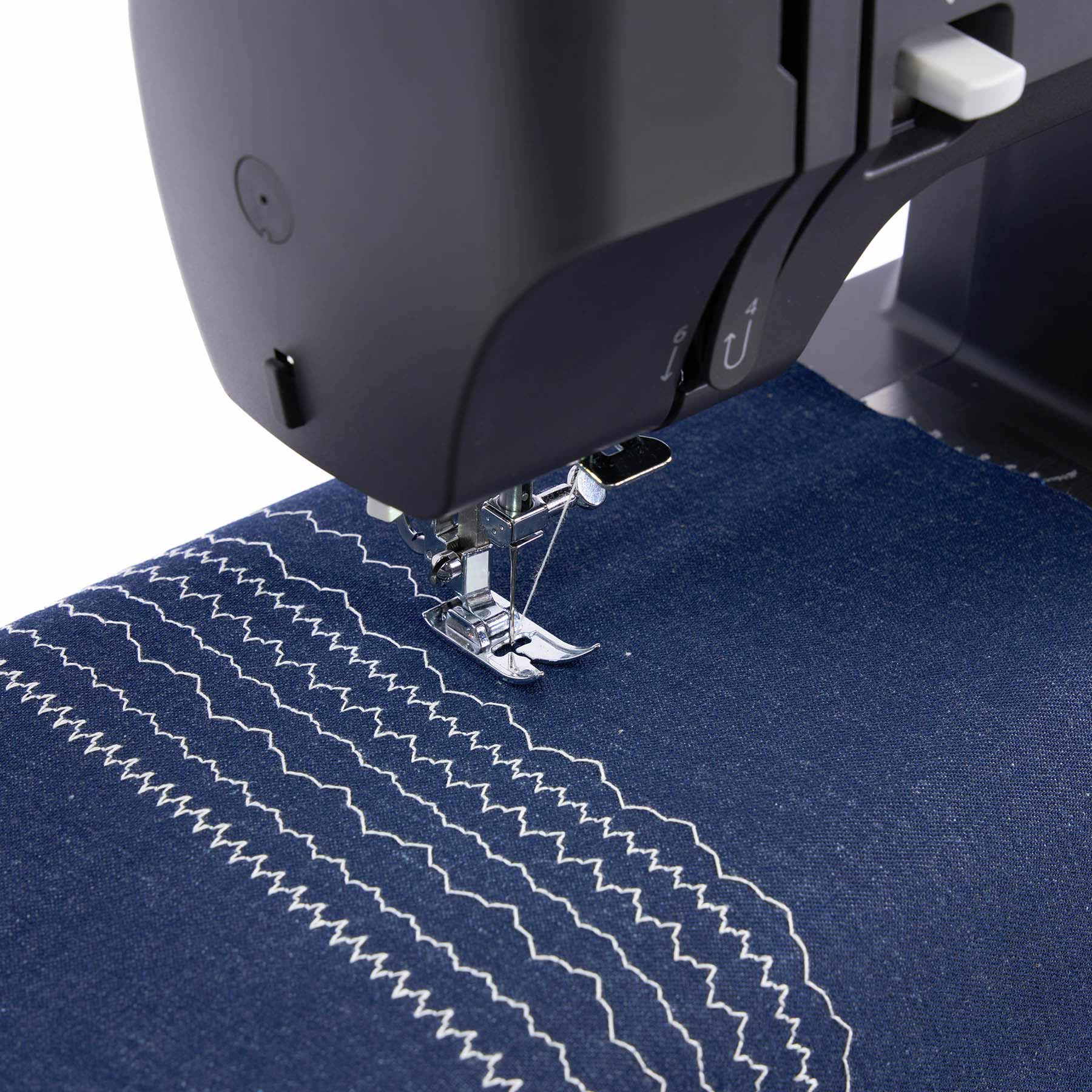 Close-up of a black sewing machine needle plate stitching dark blue fabric with white zigzag and wave stitch patterns.