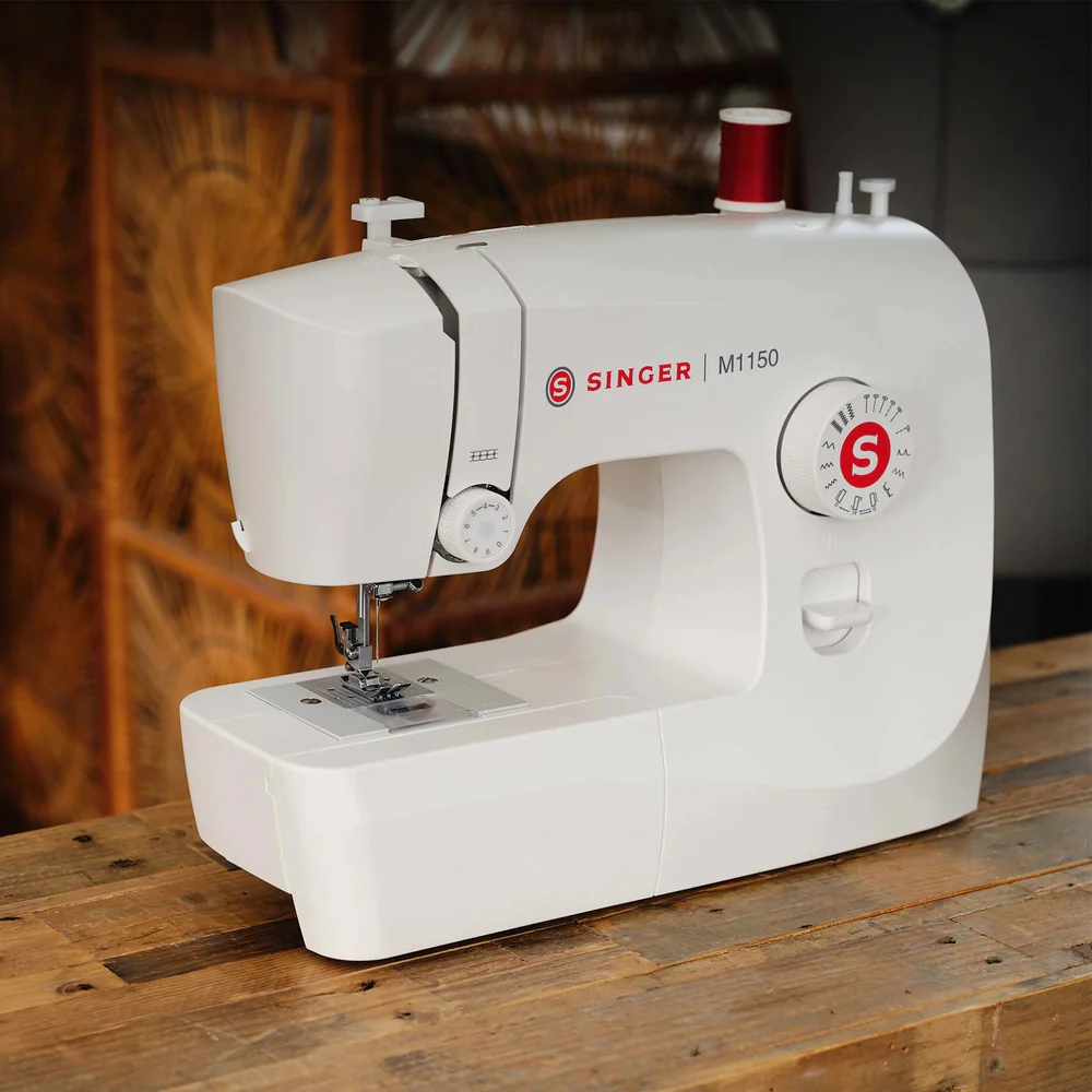 A white Singer M1150 sewing machine placed on a rustic wooden table, with a blurred warm-toned indoor background.