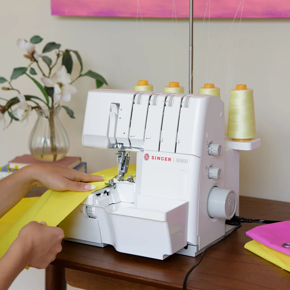 A white Singer serger sewing machine on a wooden table, with a person's hands guiding yellow fabric under the needle. Thread spools are visible on top and on the side, with a softly styled indoor background.
