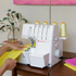 A white Singer serger sewing machine on a wooden table, with a person's hands guiding yellow fabric under the needle. Thread spools are visible on top and on the side, with a softly styled indoor background.