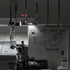 Close-up of a white serger machine showing the needle area, thread guides, presser foot, bobbin, and a printed multicolor thread guide on the front panel.