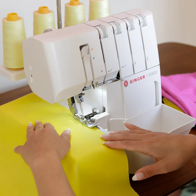 A close-up of a white Singer S0600 serger sewing machine being operated by hands guiding bright yellow fabric under the needle. Spools of thread are visible in the background, and a pink fabric piece is held on the right side.
