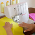 A close-up of a white Singer S0600 serger sewing machine being operated by hands guiding bright yellow fabric under the needle. Spools of thread are visible in the background, and a pink fabric piece is held on the right side.
