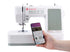 A white Singer sewing machine with a hand holding a smartphone in front of it, shown on a clean white background.
