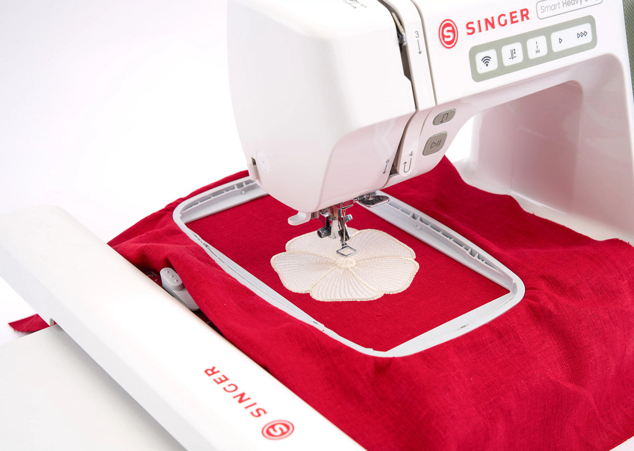 A close-up of a white Singer embroidery machine stitching a white flower-shaped fabric piece over red fabric inside a hoop, with the Singer logo visible on the machine.