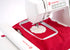 A close-up of a white Singer embroidery machine stitching a white flower-shaped fabric piece over red fabric inside a hoop, with the Singer logo visible on the machine.