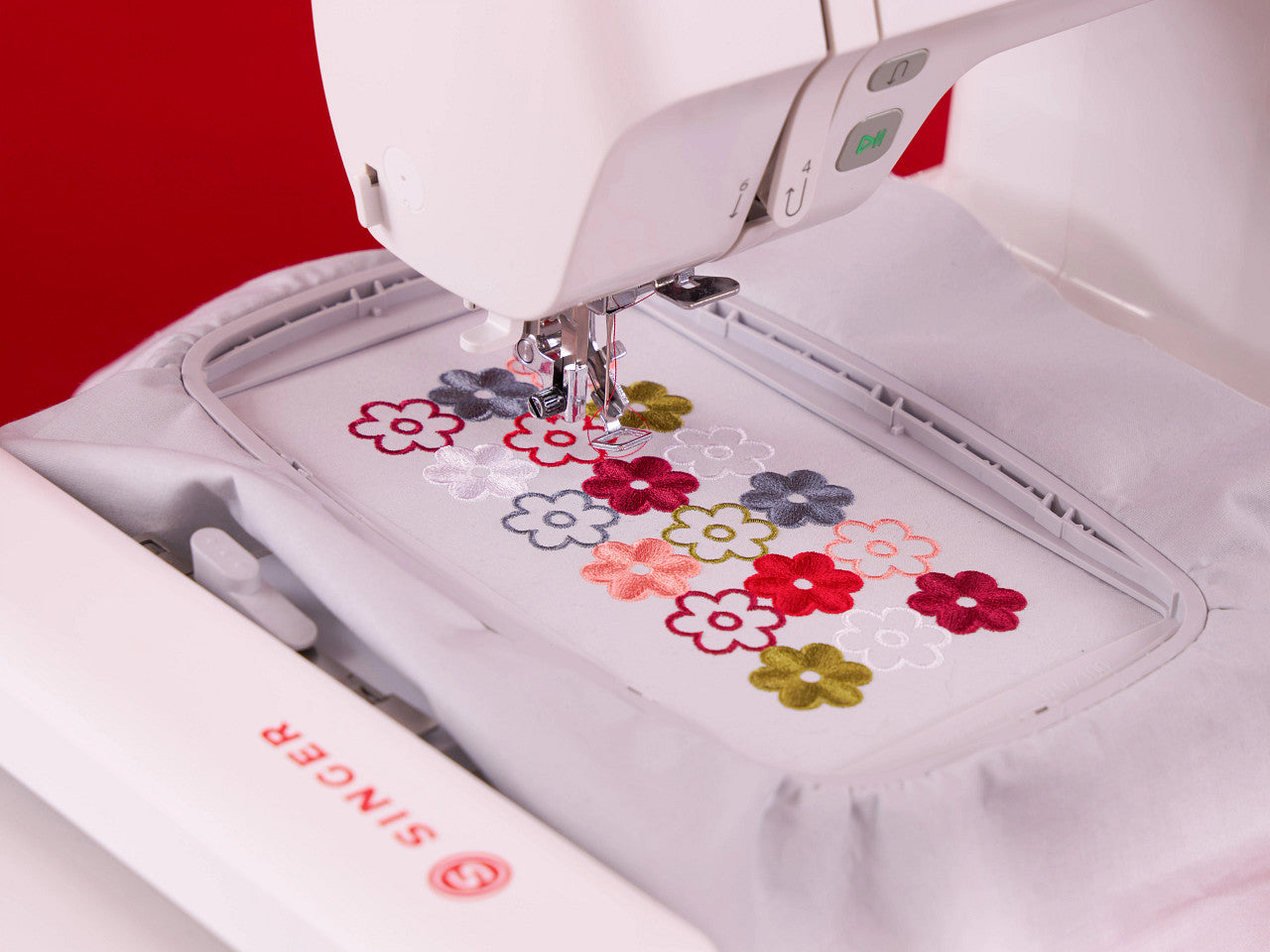 Close-up of a white Singer embroidery machine stitching colorful flower motifs into a light gray fabric underarm bag, with a red background visible behind the machine.