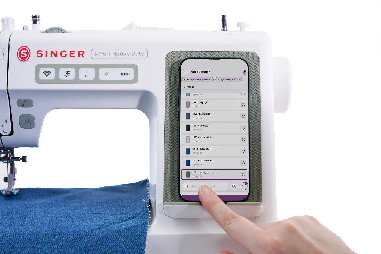 A close-up of a white Singer Smart Heavy Duty sewing machine with a person's finger touching the touchscreen interface, showing a fabric app menu. A blue piece of fabric is visible under the needle.