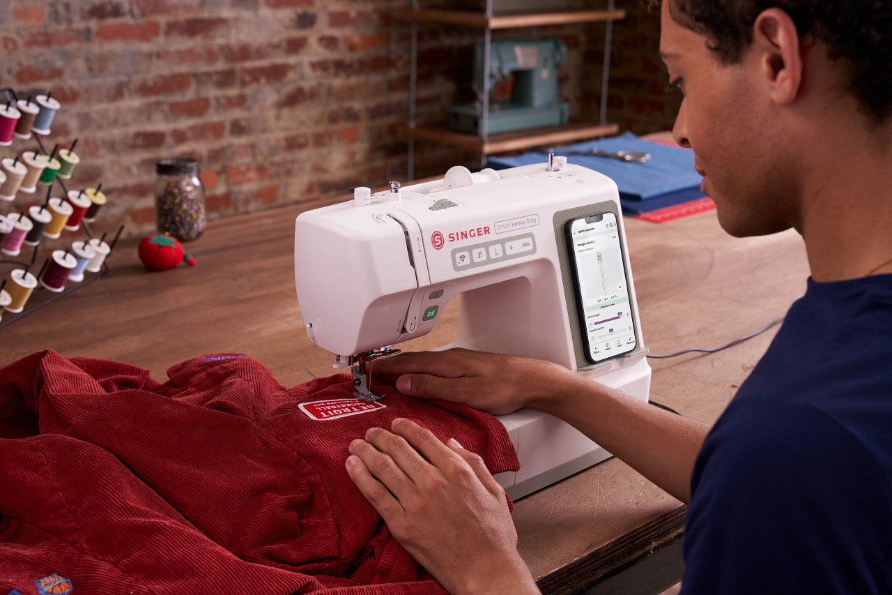 A person using a white Singer sewing machine to sew a red fabric item on a wooden table in a workshop setting.