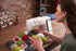 A woman operates a white Singer sewing machine on a wooden table, guiding fabric under the needle. The sewing room has a brick wall background with shelves of thread and sewing supplies.