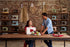 A man and a woman in a sewing studio with a brick wall and shelves of vintage sewing machines. The woman sits at a wooden table using a white sewing machine, while the man stands beside her.