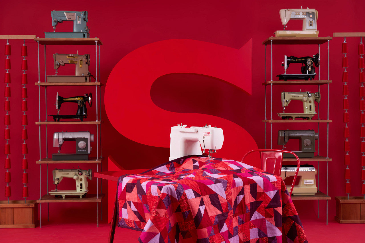 A sewing machine quilt with a geometric pattern in shades of red, pink, and purple, draped over a table in a studio setting with a large red background and shelves of vintage sewing machines.
