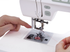Close-up of a white sewing machine with a person's hands adjusting the needle area and feeding thread, shown against a clean white background.