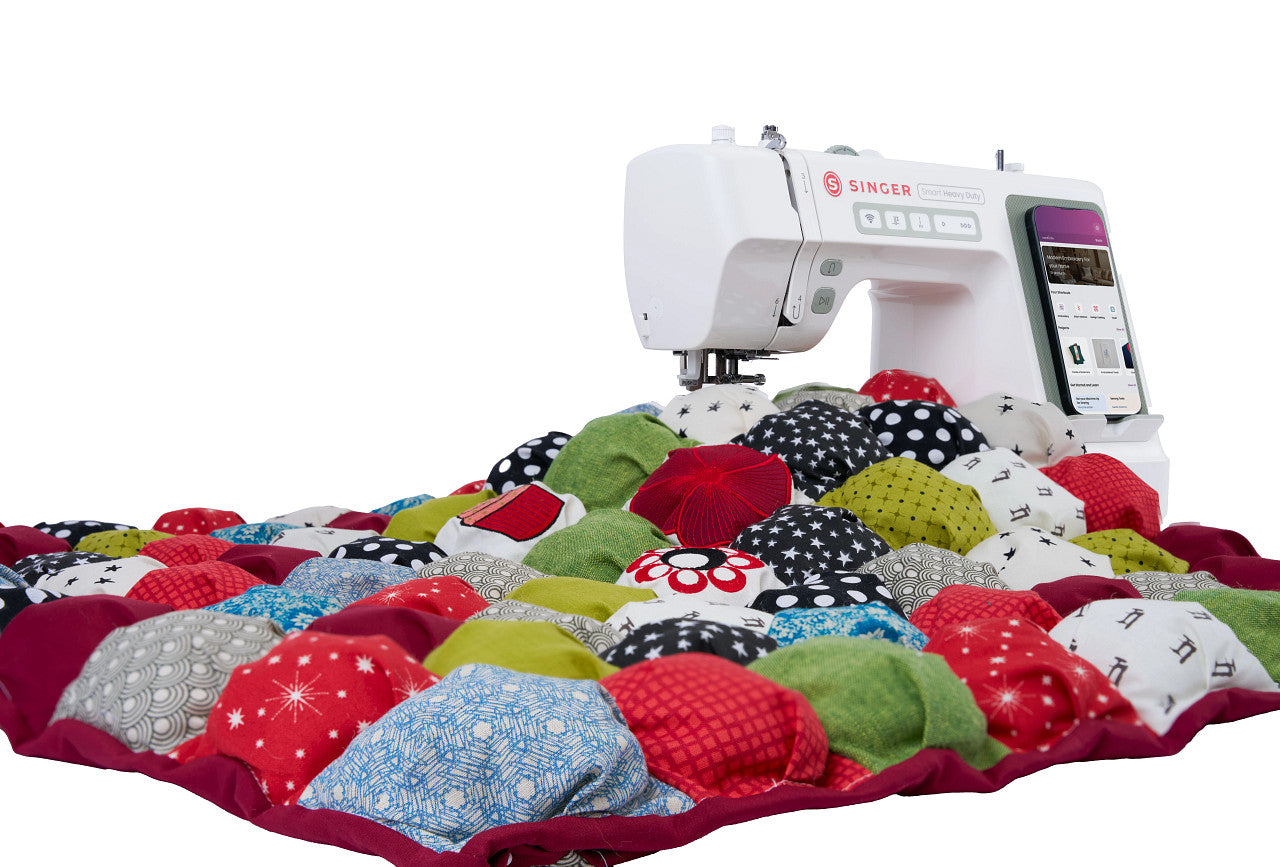 A white Singer embroidery machine positioned over a colorful patchwork quilt made of many fabric squares in red, green, blue, black, white, and yellow, photographed on a clean white background.